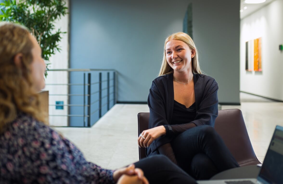 Celina sidder på sofastol og snakker smilende til anden medarbejder